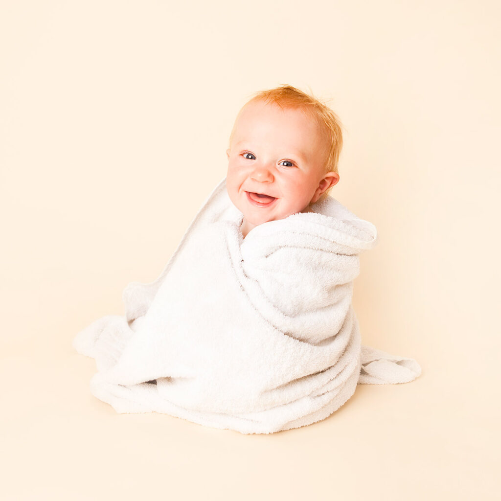 Cute smiley baby sitting on Hertford studio floor while a white towel is wrapped around him.  