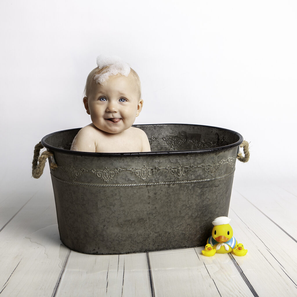 Baby girl is sitting in a metal bath tub having her tongue out.