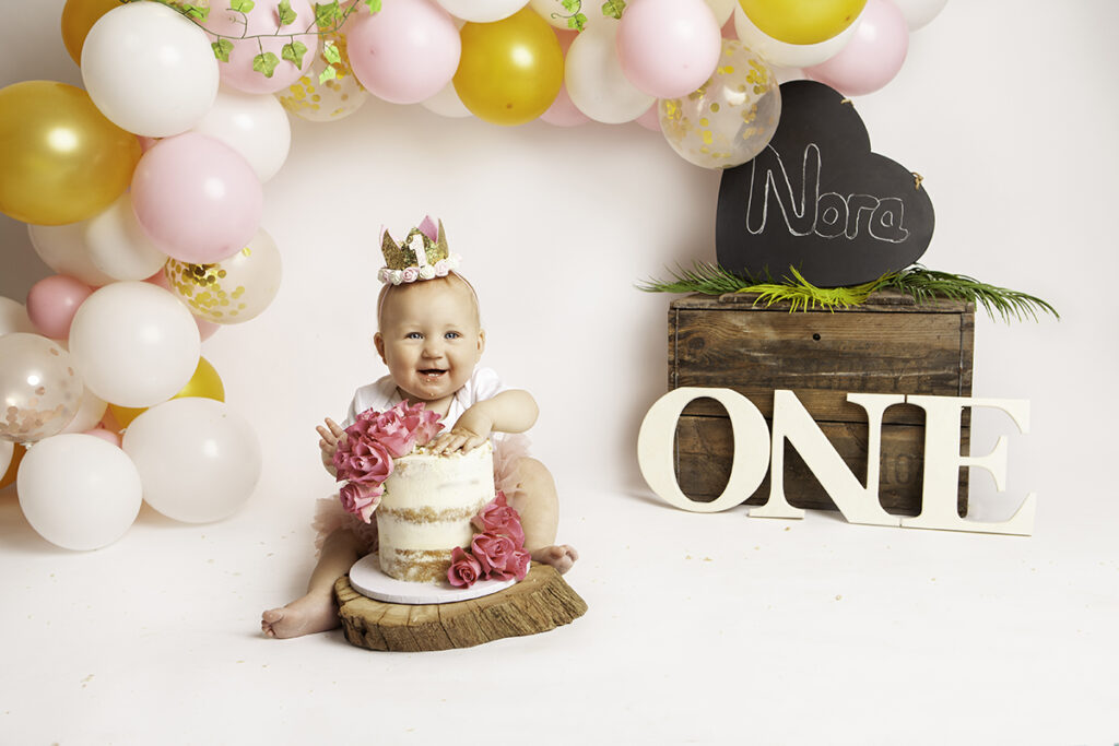 Nora's first birthday. Baby girls is sitting behind her cake with colourful balloons in the background.
