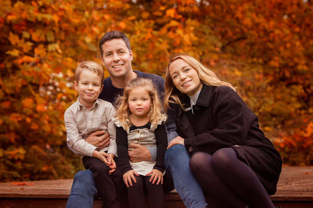 A family of 4 sitting on a wooden deck smiling to the camera. You can see autumn leaves in the background. 