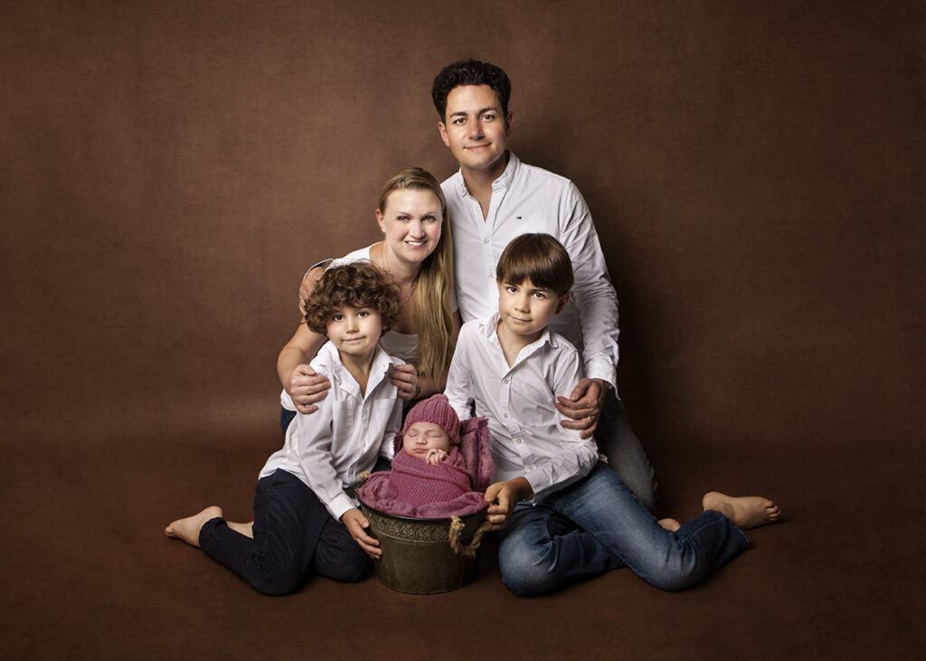 A family of five photograph where every family member has a white shirt on. Newborn baby girl is in a basket nicely wrapped  up with a hat on.