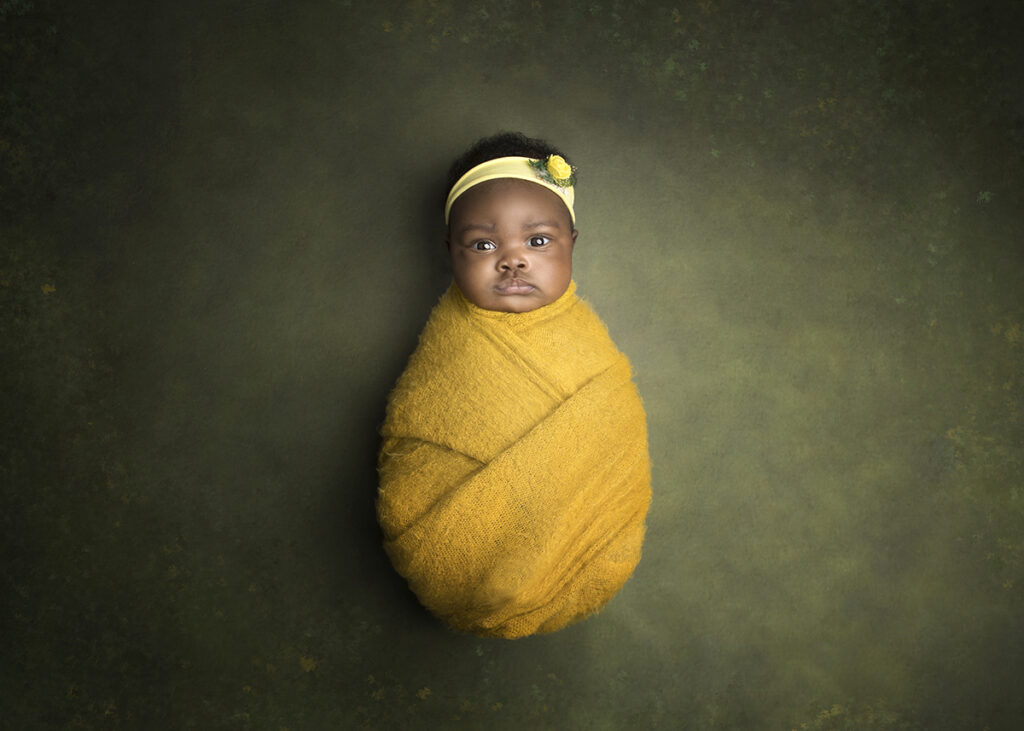 Baby girl swaddled in a bright yellow wrap wearing a headpiece with yellow rose during her newborn photo shoot. 