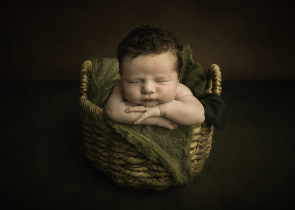 Baby boy leaning out from a basket with his arms under his chin. The newborn has lots of brown hair.