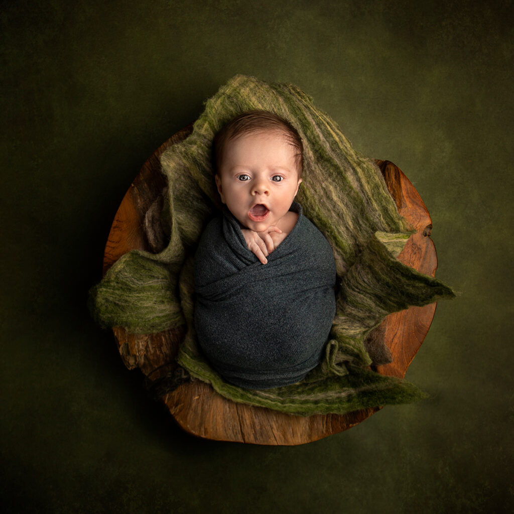 There is a newborn baby swaddled in a fruit basket. He is looking into the camera while yawning. His parants are behind the camera.