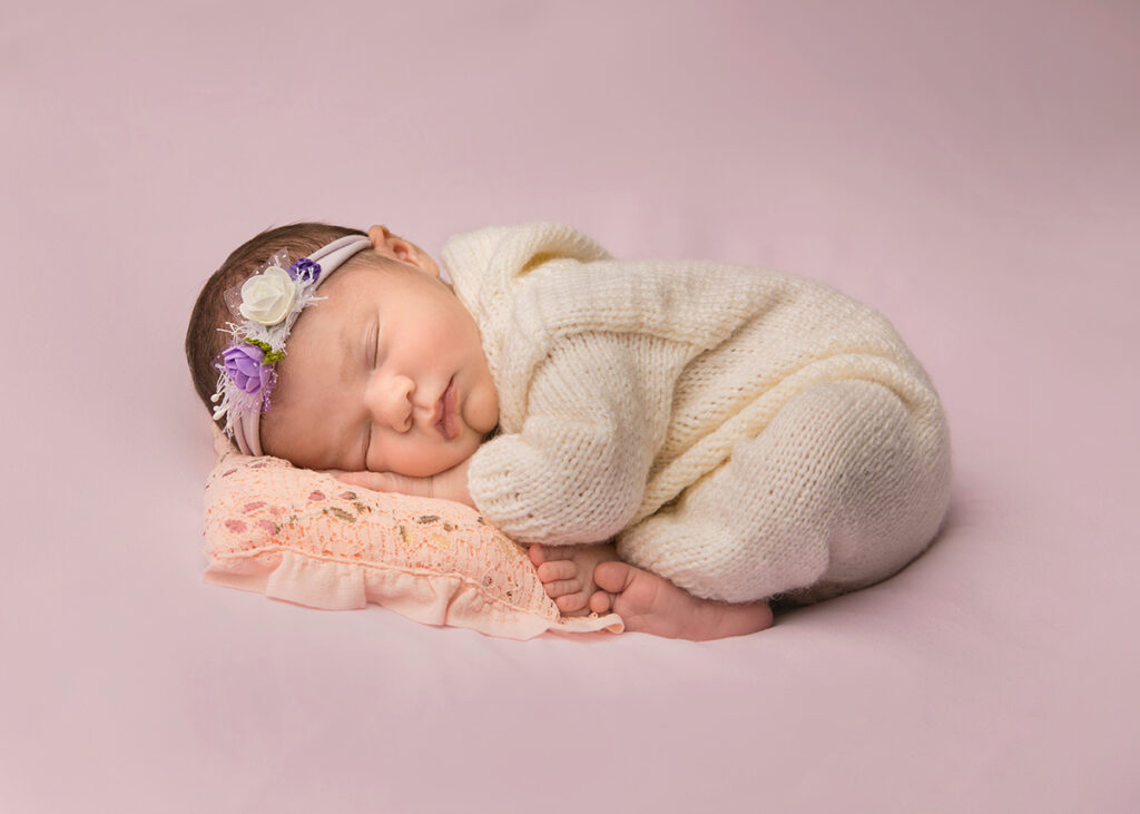 Newborn girl sleeping in womb position on a beanbag. She is wearing white knited onesie with a hood.