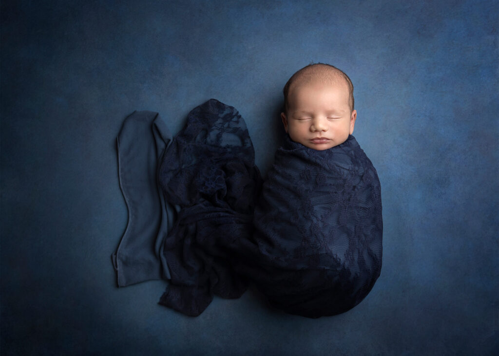 Newborn swaddled in blue wrap. He is sleeping on a blue backdrop in the studio.